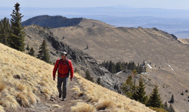 A male hiker in a red jacket hikes up steep alpine trail with a mountain ridge in the background